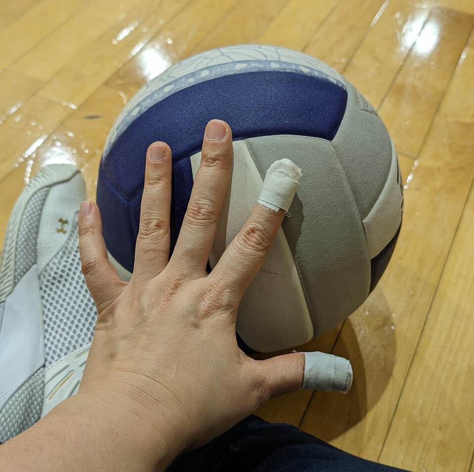 A hand palming a volleyball on a wooden floor with her left index and thumb taped up.